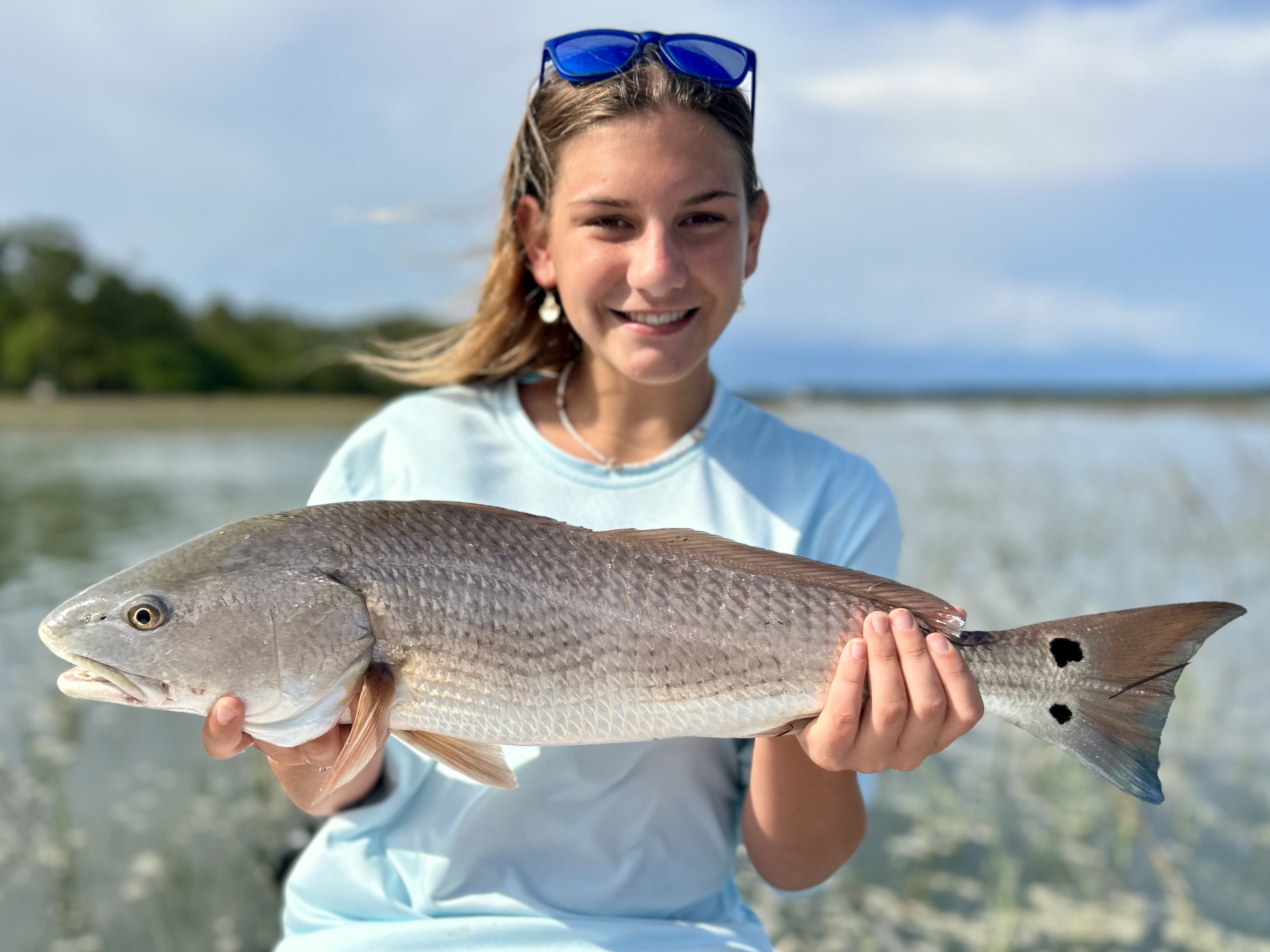A young girl proudly holds a redfish while standing by the water in Beaufort, South Carolina. She is wearing a light blue shirt and sunglasses, with a shoreline and greenery in the background.