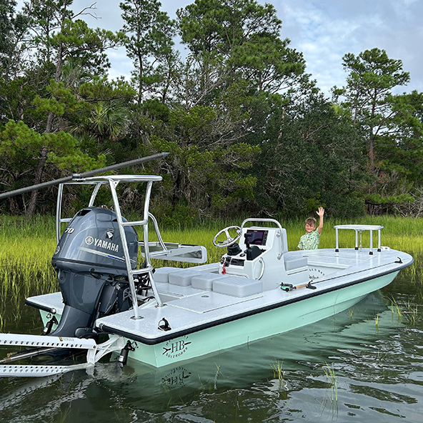 A boy waving from a small boat with an outboard Yamaha motor, surrounded by tall green grass and trees.