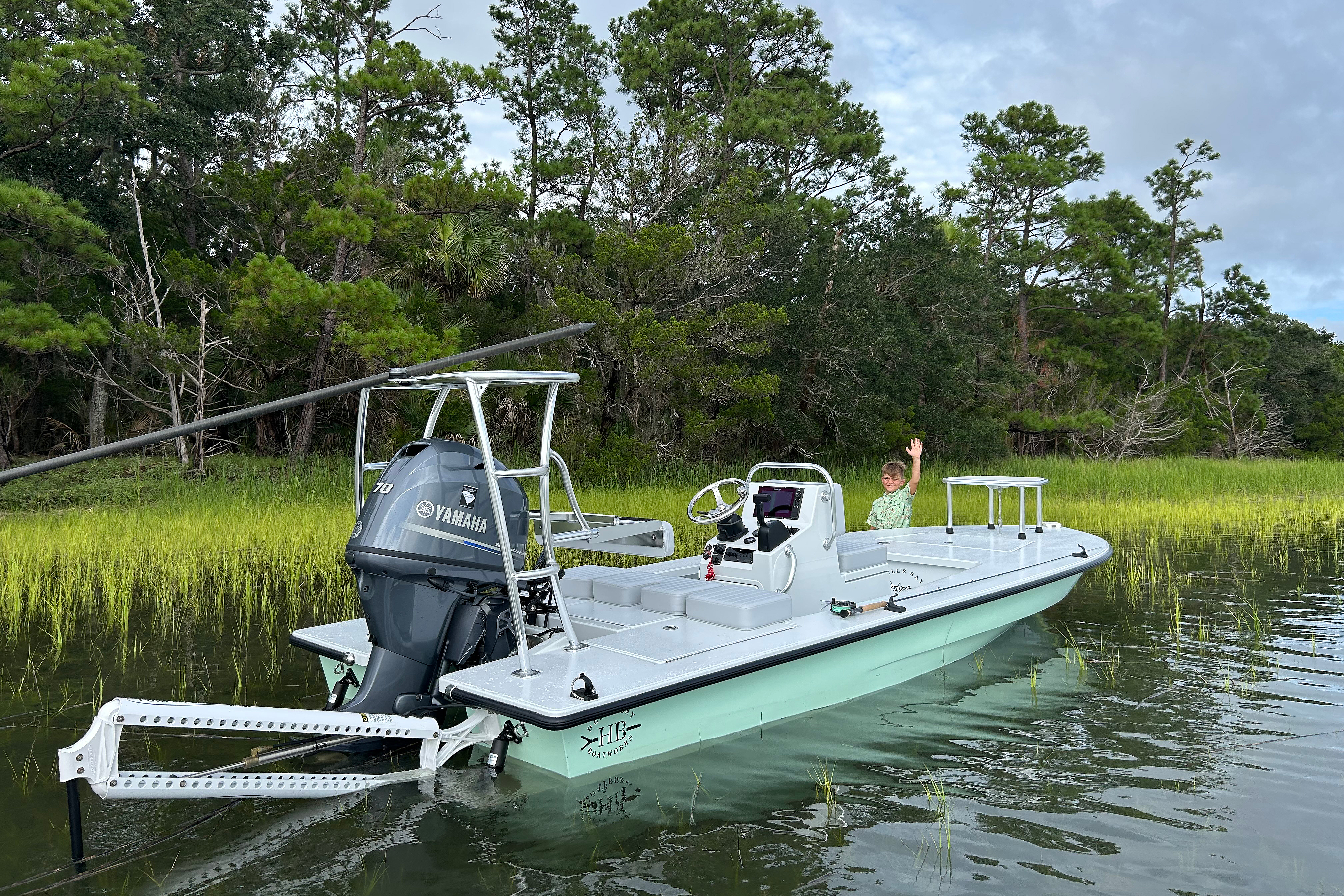 A child waving from a fishing skiff on a calm waterway surrounded by tall grass and trees.