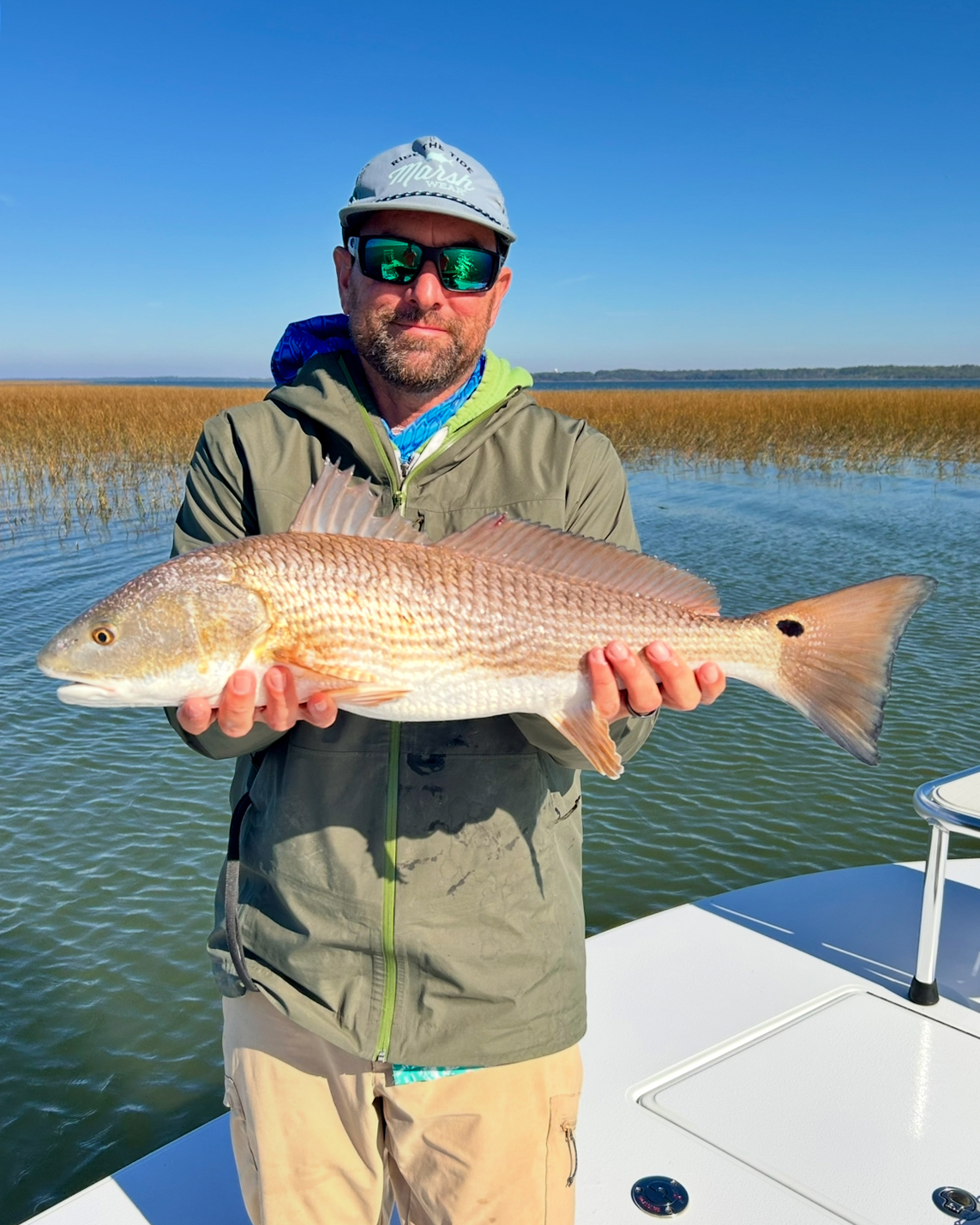 A person holding a redfish while standing on a boat, with marsh grass and water visible in the background.