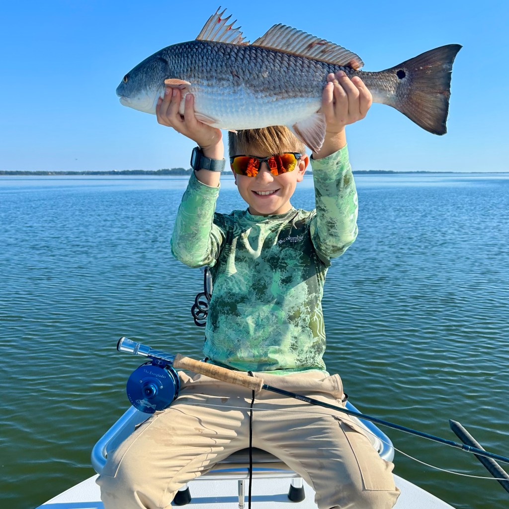 A young boy wearing sunglasses proudly holds up a redfish while sitting on a boat, with calm water and a clear blue sky in the background.