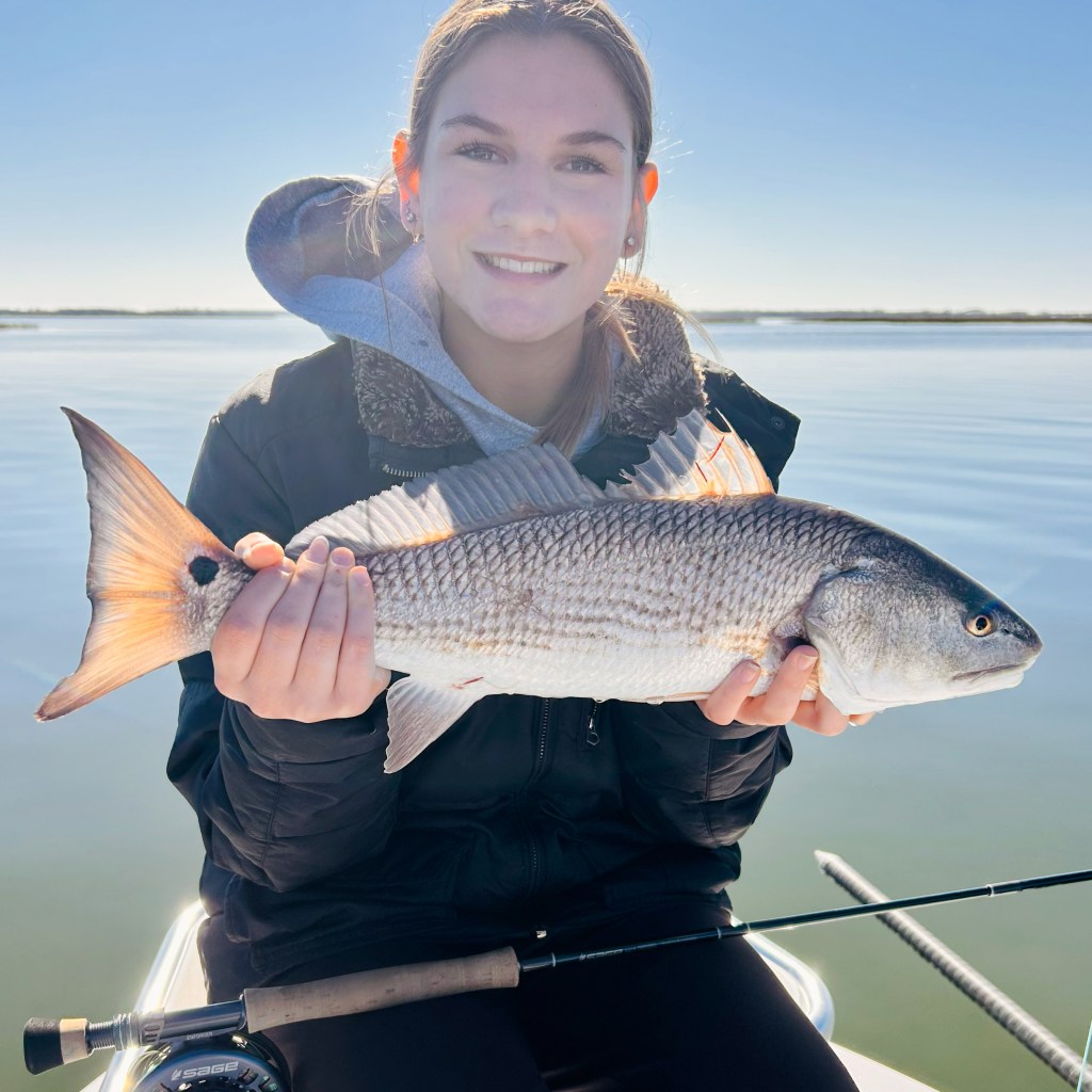 A young person holding a redfish while smiling on a boat, with calm water and a clear sky in the background.