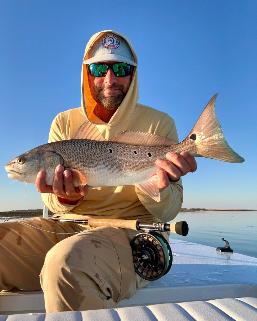 A person holds a redfish while sitting on a fishing skiff, wearing sunglasses and a hat, under a clear blue sky.